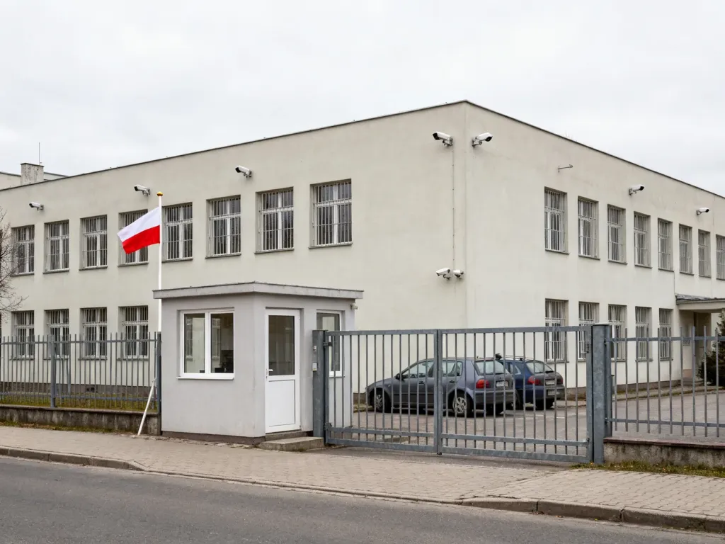 Wejście do Aresztu Śledczego w Gdańsku z bramą, budką strażniczą, ogrodzeniem i budynkiem z oknami z kratami.